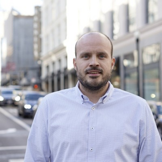 Man facing camera, in a dress shirt, city street behind him, with a close-cropped dark beard beard, light skin, and very short hair.