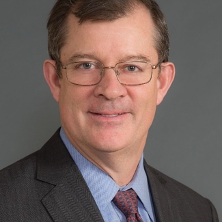 A slightly smiling man with short brown hair, white skin, and thin-rimmed glasses.