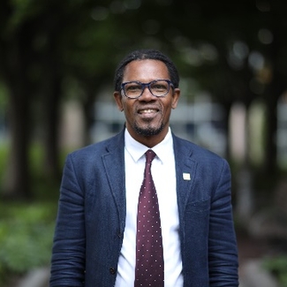 A man looks directly into the camera, smiling, with closs-cropped beard and hair, dark brown skin, black-rimmed glasses, wearing a suit and tie, with out-of focus city park behind.